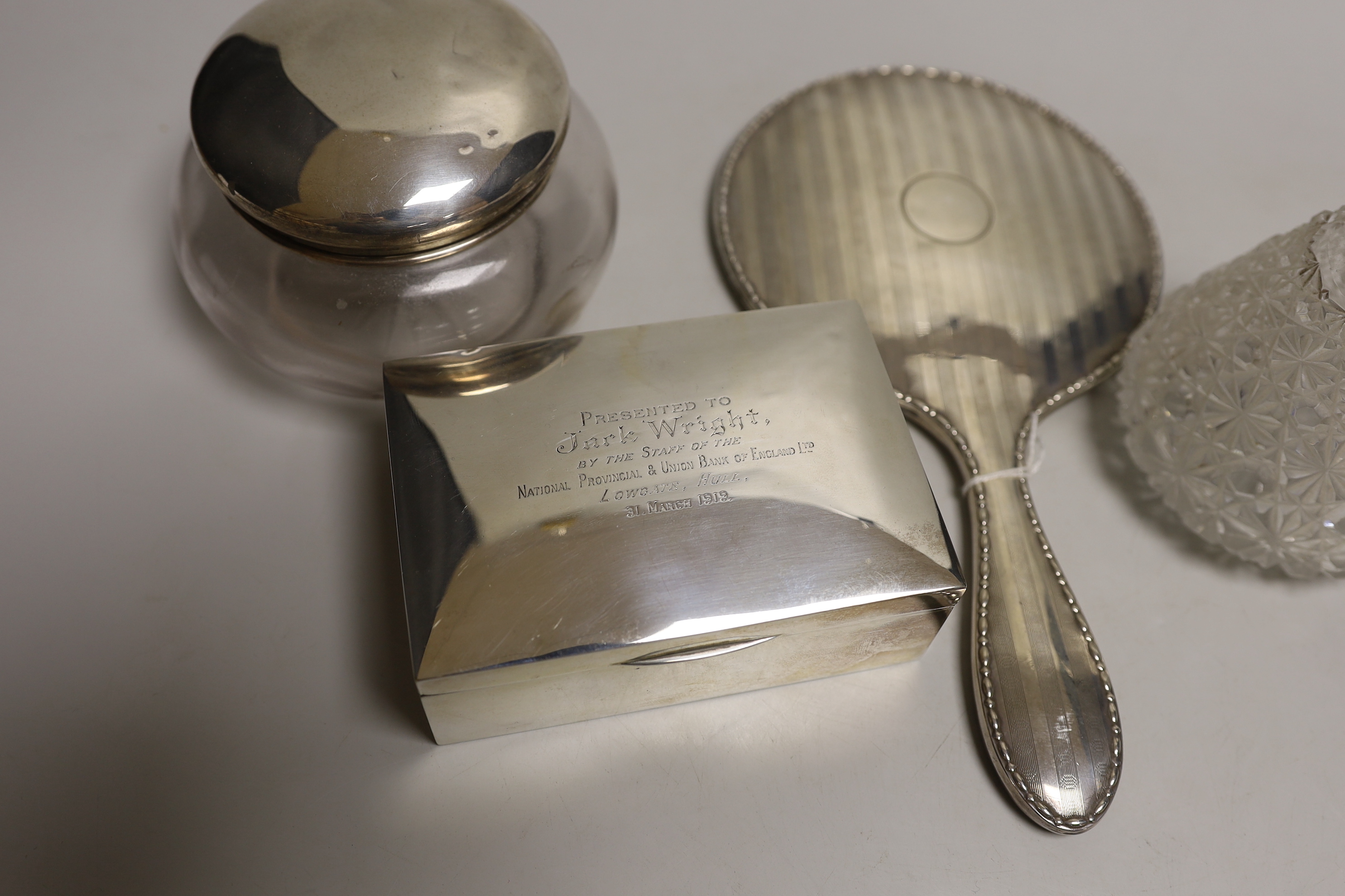 A George V silver backed hand mirror, a silver mounted cigarette box, a silver topped powder bowl and a silver topped scent bottle.